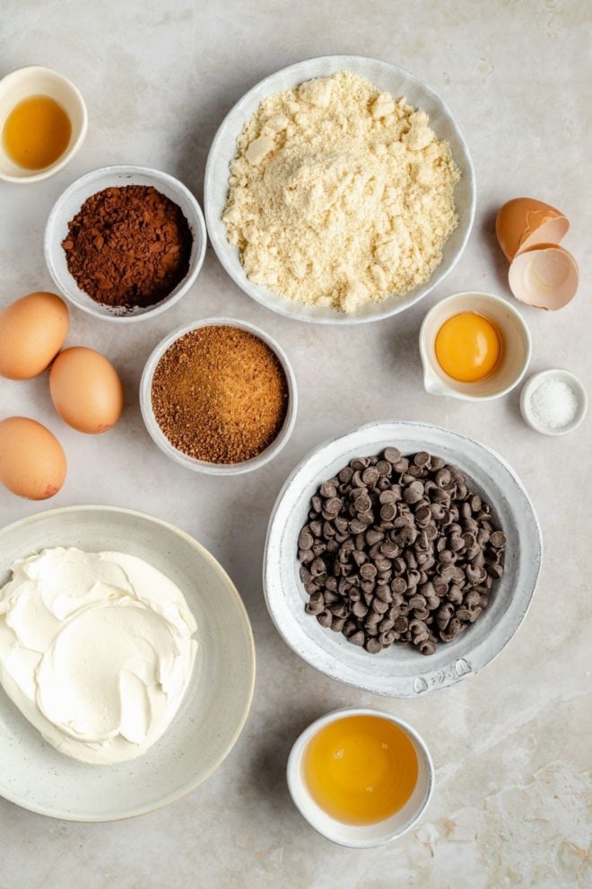 This is an overhead image of ingredients laid out on a counter. The ingredients are almond flour, cocoa powder, coconut sugar, chocolate chips, eggs, and cream cheese.