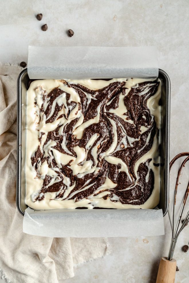 This is an overhead image of a square pan with a piece of parchment paper draping over two sides. Raw brownie batter with a cream cheese swirl is inside the pan. The pan sits on a white and grey counter. A tan towel is in the bottom left corner of the image. A whisk with raw brownie batter is in the bottom right of the image. Some chocolate chips are scattered above the pan to the top left of the image.