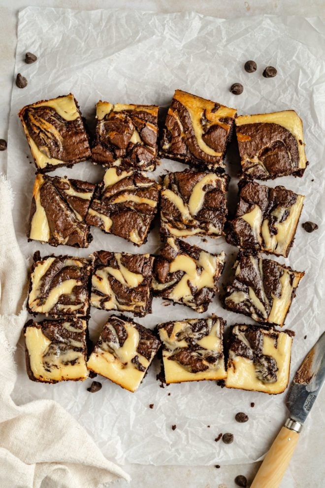 This is an overhead image of brownies with a cheesecake swirl. The brownies are cut into squares and sit on a white piece of parchment paper on a white counter. A knife is in the bottom corner of the image.