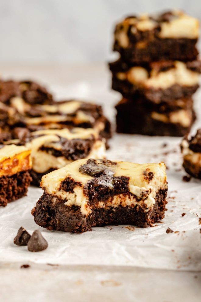 This is a side image of a brownie with a cheesecake top layer and a bite taken out. The brownie sits on a white piece of parchment paper and more brownies are blurred in the background.