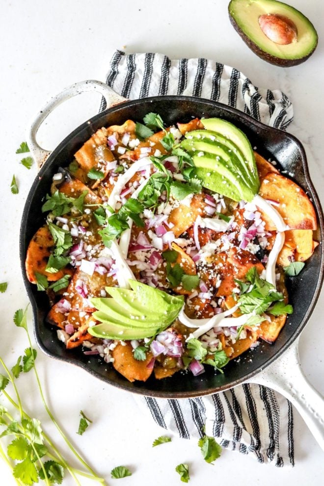 This is an overhead image of chilaquiles verdes in a white skillet on a white counter on top of a black and white striped towel. The chilaquiles are topped with avocado, cotija cheese, cilantro, and red onion. More cilantro leaves and half of an avocado are on the counter next to the skillet.