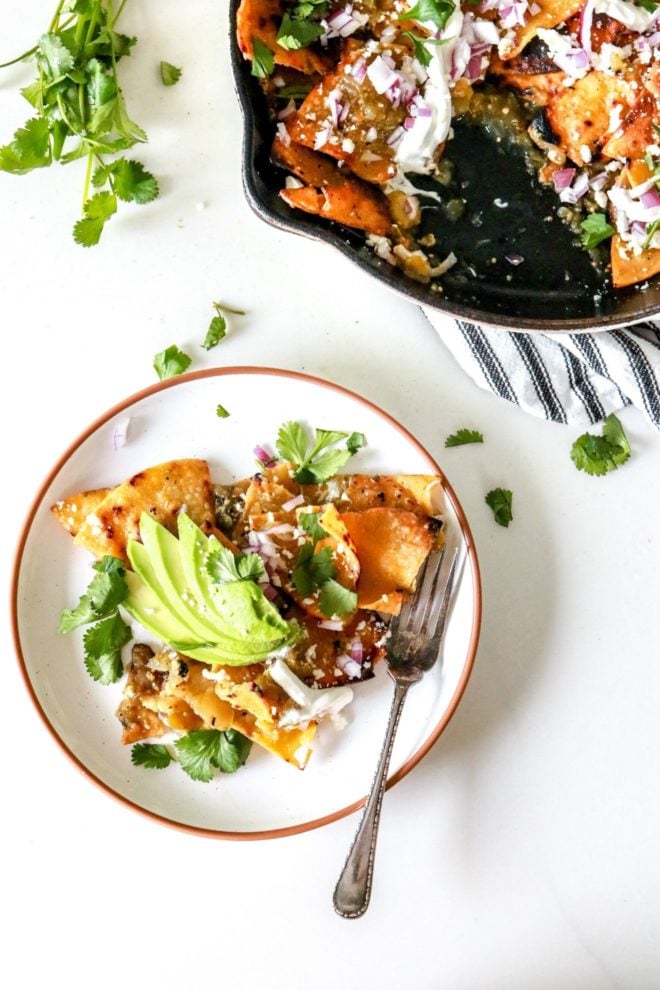 This is an overhead image of a plate on a white counter. One the plate are chilaquiles verdes topped with avocado, cilantro, cotija, and onion. A fork lays on the side of the dish. A skillet with more chilaquiles verdes is in the top right corner of the image. More cilantro leaves are on the counter toward the top of the plate and the top left of the image.