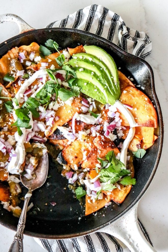 This is an overhead image closeup of chilaquiles verdes in a white skillet on a white counter on top of a black and white striped towel. The chilaquiles are topped with avocado, cotija cheese, cilantro, and red onion.