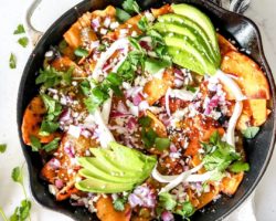 This is an overhead image of chilaquiles verdes in a white skillet on a white counter on top of a black and white striped towel. The chilaquiles are topped with avocado, cotija cheese, cilantro, and red onion. More cilantro leaves are on the counter next to the skillet.
