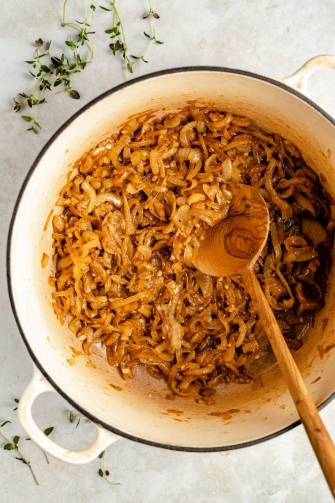 This is an overhead image of caramelized onions in a white pot. A wooden spoon is sticking out of the white pot. The pot sits on a white counter with fresh thyme leaves around it.