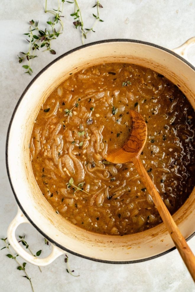 This is an overhead image of a large pot with broth, caramelized onions, and a wooden spoon. The pot sits on a white counter with thyme leaves around it.