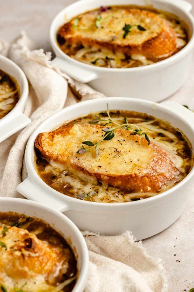 This is a side image of four white crocks filled with french onion soup. The soup is topped with French bread and Gruyere cheese and a thyme sprig. The crocks sit on a white counter and an ivory tea towel is weaved in between the crocks.