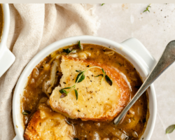 This is an overhead image of a white crock filled with french onion soup. On top of the soup is a piece of French bread, melted cheese, and thyme. The crock sits on a white counter with an off-white tea towel to the left side of the image. Text overlay reads "vegetarian French onion soup."
