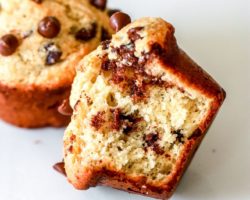 This is a mini muffin with mini chocolate chips. A bite is taken out of the muffin and it is laying on its side on a white counter. Another muffin is blurred in the background.
