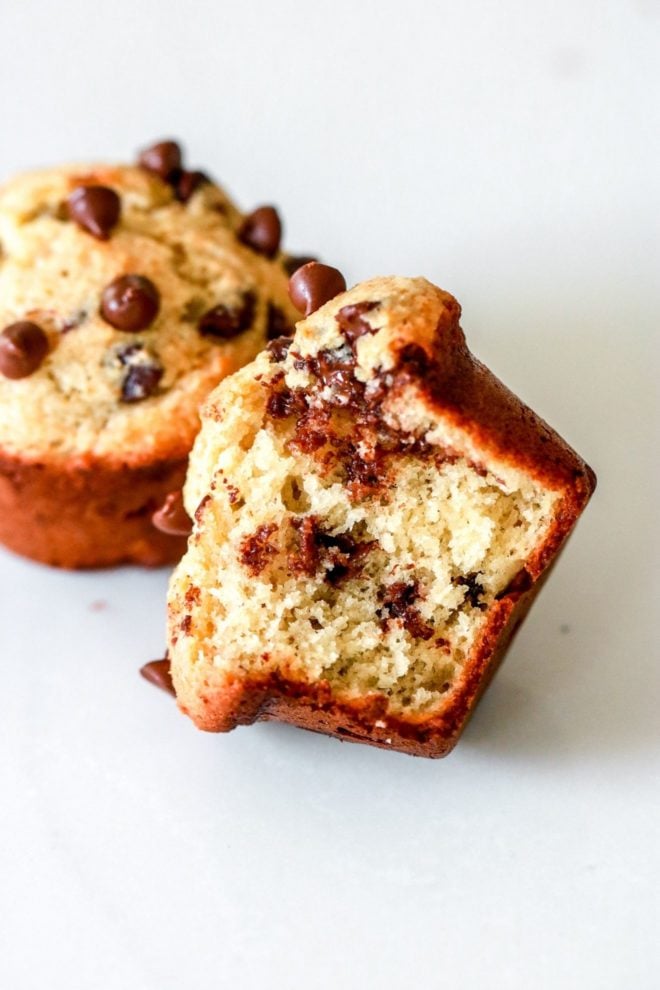 This is a mini muffin with mini chocolate chips. A bite is taken out of the muffin and it is laying on its side on a white counter. Another muffin is blurred in the background.