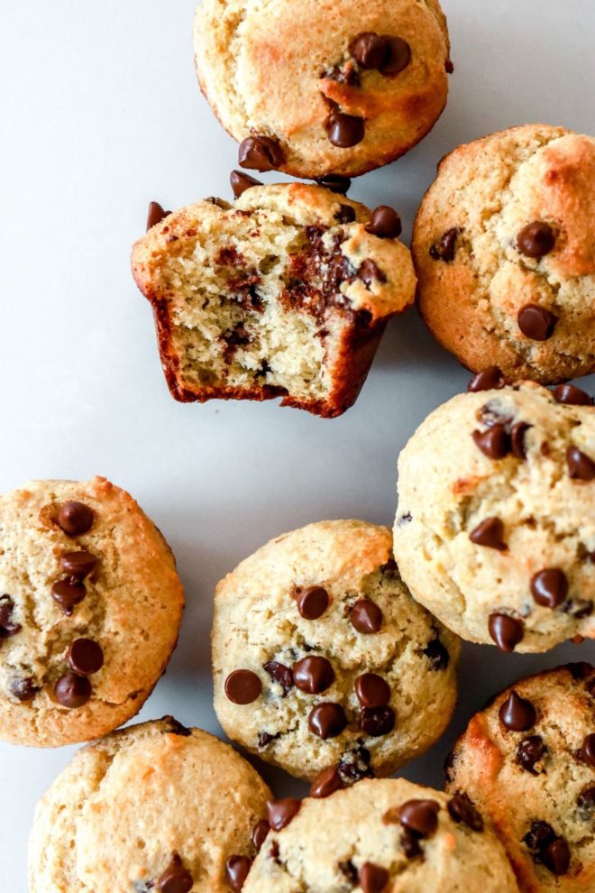 This is an overhead image of a bunch of mini muffins all next to each other. The muffins have mini chocolate chips in them and on top. One muffin has a bite taken out and is laying on its side.