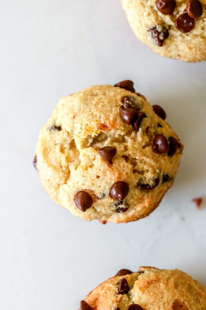 This is an overhead image of the top of a muffin with mini chocolate chips on top. The muffin sits on a white counter. Two more chocolate chip muffins are in the top right and bottom right of the image.