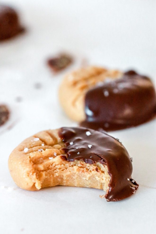 This is a side view of a peanut butter cookie half dipped in chocolate. The cookie is sprinkled with salt and has a bite taken out of it. Another cookie is blurred in the background. Both cookies sit on a white counter.