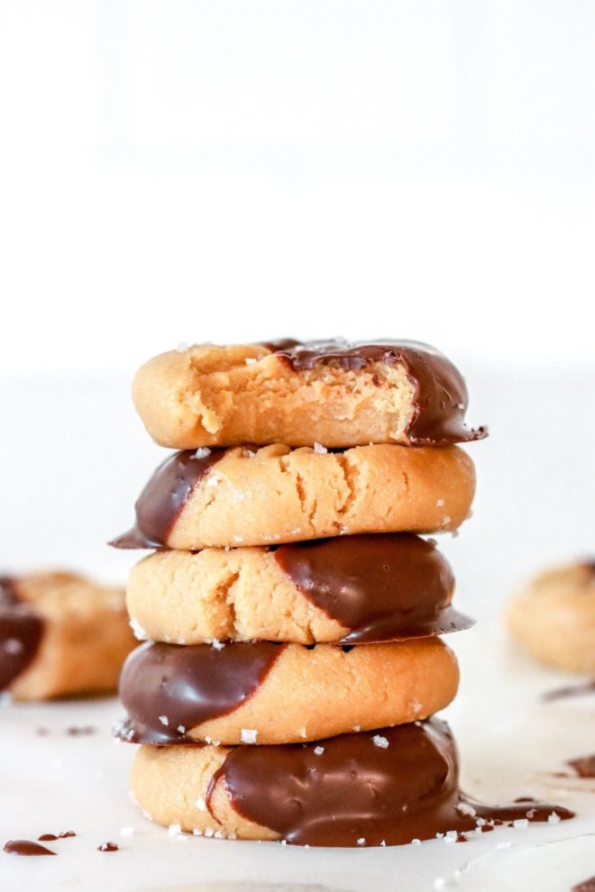 This is a stack of peanut butter cookies sitting on a white counter. The cookies are half dipped in chocolate and the top cookie has a bite taken out. More cookies are blurred in the background.