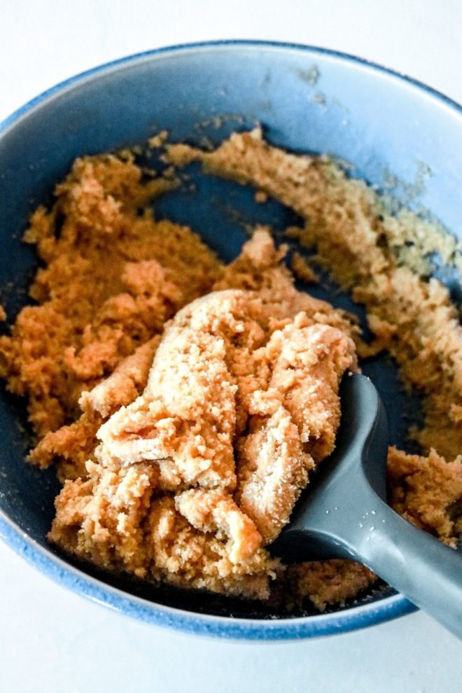 This is a blue bowl with peanut butter cookie dough in it. The bowl sits on a white counter and a rubber spatula scoops in the bowl and leans against the side.
