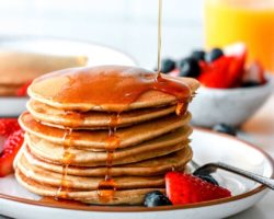 This is a side view of a stack of pancakes on a plate with berries. Syrup is being poured on top of the pancakes. More berries, pancakes, and orange juice is blurred in the background.