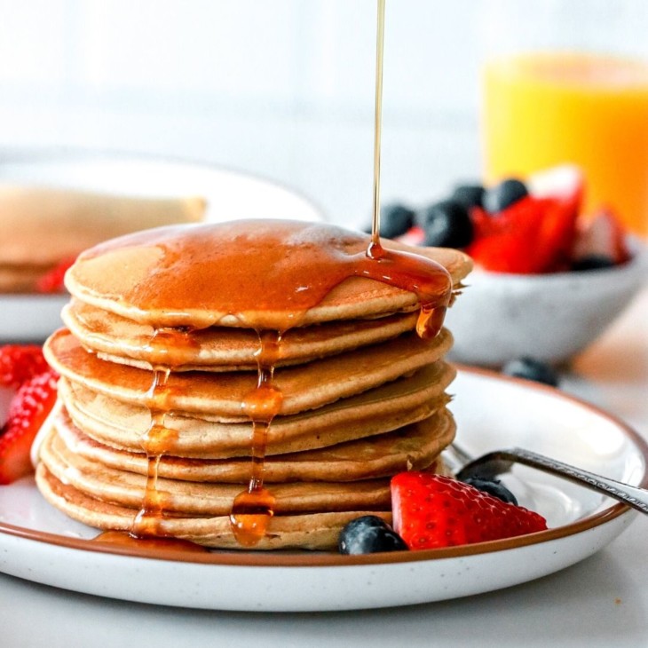 This is a side view of a stack of pancakes on a plate with berries. Syrup is being poured on top of the pancakes. More berries, pancakes, and orange juice is blurred in the background.