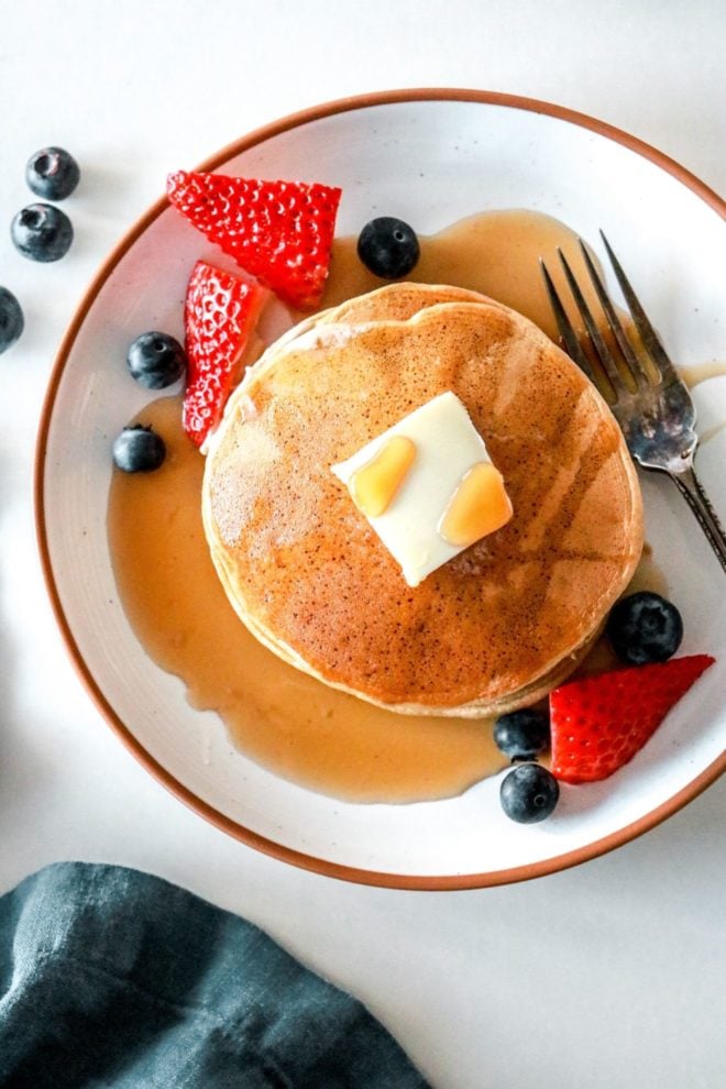 This is an overhead image of pancakes on a plate with strawberries and blueberries. The pancakes have a pad of butter and syrup on them.