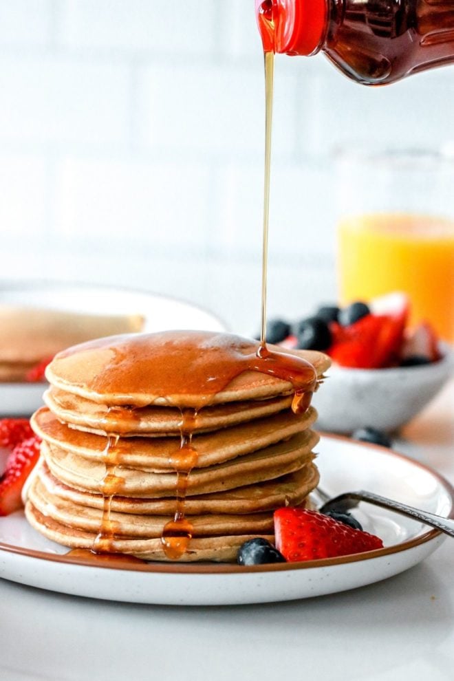 This is a side view of a stack of oat flour pancakes on a plate with berries. Syrup is being poured on top of the pancakes. More berries, pancakes, and orange juice is blurred in the background.