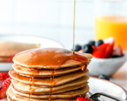 This is a side view of a stack of pancakes on a plate with berries. Syrup is being poured on top of the pancakes. More berries, pancakes, and orange juice is blurred in the background. Text overlay reads "healthy oat flour pancakes"