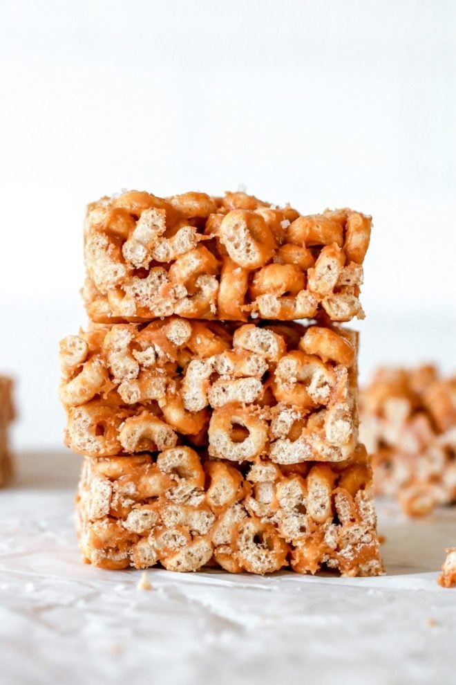 This is a side view of three cheerio bars stacked top of each other. The bars are sitting on a white surface and a white background. More cereal bars are blurred in the background.
