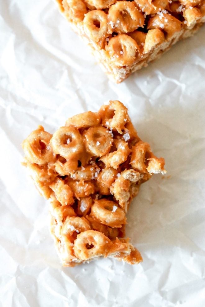 This is an overhead image of a peanut butter cheerio bar. The bar is in the center bottom of the image and has a bite taken out. The bar sits on a white piece of parchment paper and another cheerio bar is on the top of the image.