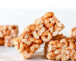 This is a side view of a cheerio bar leaning against another bar. More cereal bars are blurred in the background. The bars sit on a white surface with a white background.