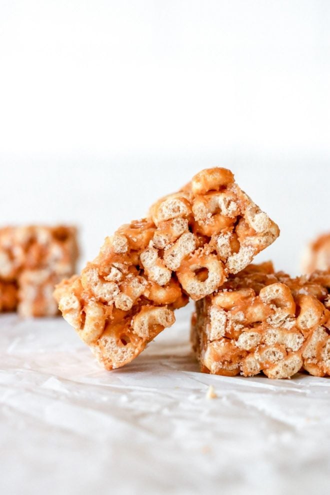 This is a side view of a cheerio bar leaning against another bar. More cereal bars are blurred in the background. The bars sit on a white surface with a white background.