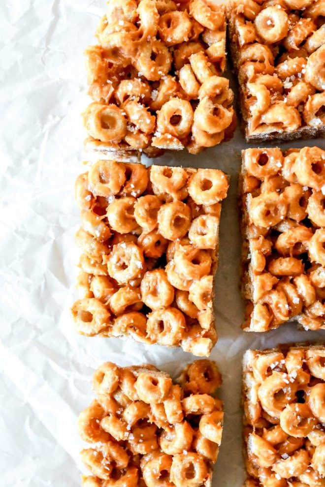 This is an overhead image of peanut butter cheerio bars. The bars are cut and sprinkled in salt. The bars sit on a white piece of parchment paper.
