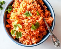 This is an overhead image of a blue bowl filled with Spanish cauliflower rice. The rice is a deep orange color and topped with fresh cilantro leaves. More cilantro leaves are on the counter, surrounding the bowl. An antique spoon is scooping some rice from the bowl and leans against the side of the bowl.