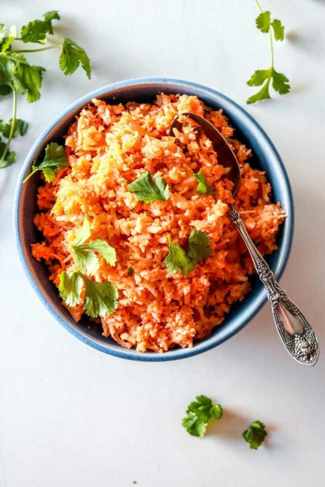This is an overhead image of a blue bowl filled with Spanish cauliflower rice. The rice is a deep orange color and topped with fresh cilantro leaves. More cilantro leaves are on the counter, surrounding the bowl. An antique spoon is scooping some rice from the bowl and leans against the side of the bowl. 