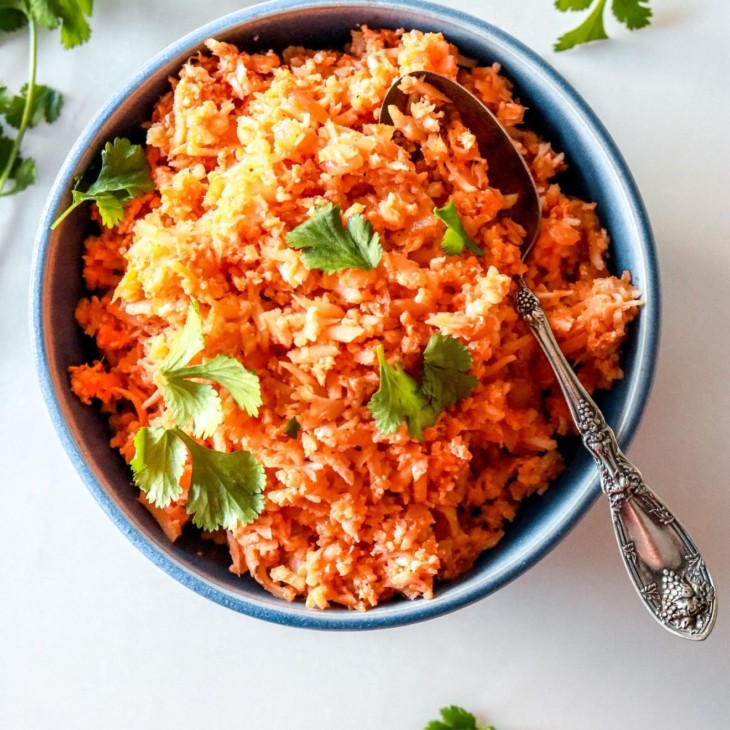 This is an overhead image of a blue bowl filled with Spanish cauliflower rice. The rice is a deep orange color and topped with fresh cilantro leaves. More cilantro leaves are on the counter, surrounding the bowl. An antique spoon is scooping some rice from the bowl and leans against the side of the bowl.