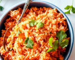 This is an overhead image of a blue bowl filled with Spanish cauliflower rice. The rice is a deep orange color and topped with fresh cilantro leaves. More cilantro leaves are on the counter, surrounding the bowl. An antique spoon is scooping some rice from the bowl and leans against the side of the bowl. Text overlay reads "spanish cauliflower rice keto & gluten free"