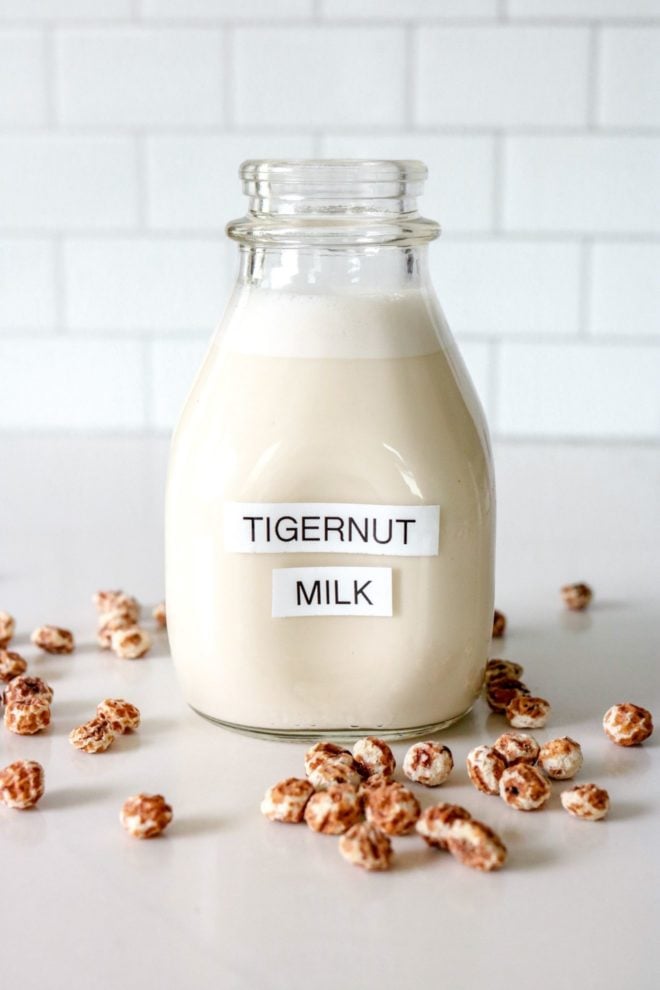This is a side view of a glass milk bottle on a white marble counter with white tile in the background. Inside the glass container is a white milk substance. The bottle is labeled "tigernut milk" and there are tigernuts scattered on the counter around the bottle.