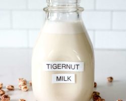 This is a side view of a glass milk bottle on a white marble counter with white tile in the background. Inside the glass container is a white milk substance. The bottle is labeled "tigernut milk" and there are tigernuts scattered on the counter around the bottle.
