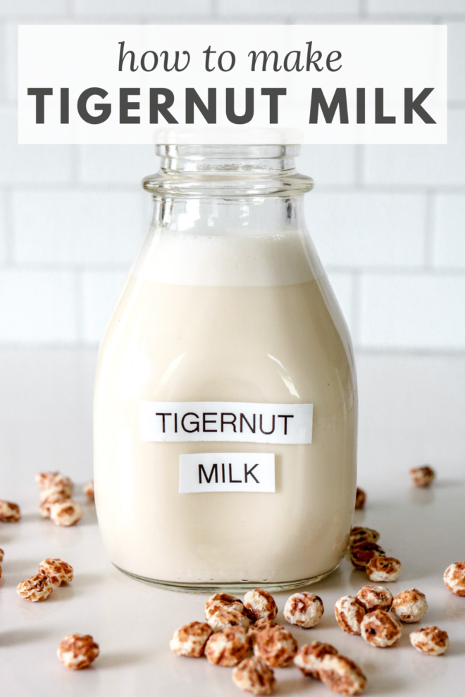 This is a side view of a glass milk bottle on a white marble counter with white tile in the background. Inside the glass container is a white milk substance. The bottle is labeled "tigernut milk" and there are tigernuts scattered on the counter around the bottle.