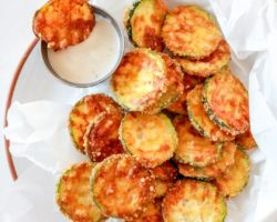 This is an overhead image of a plate of zucchini chips on top of parchment paper. There is a small bowl of ranch dressing to the top.