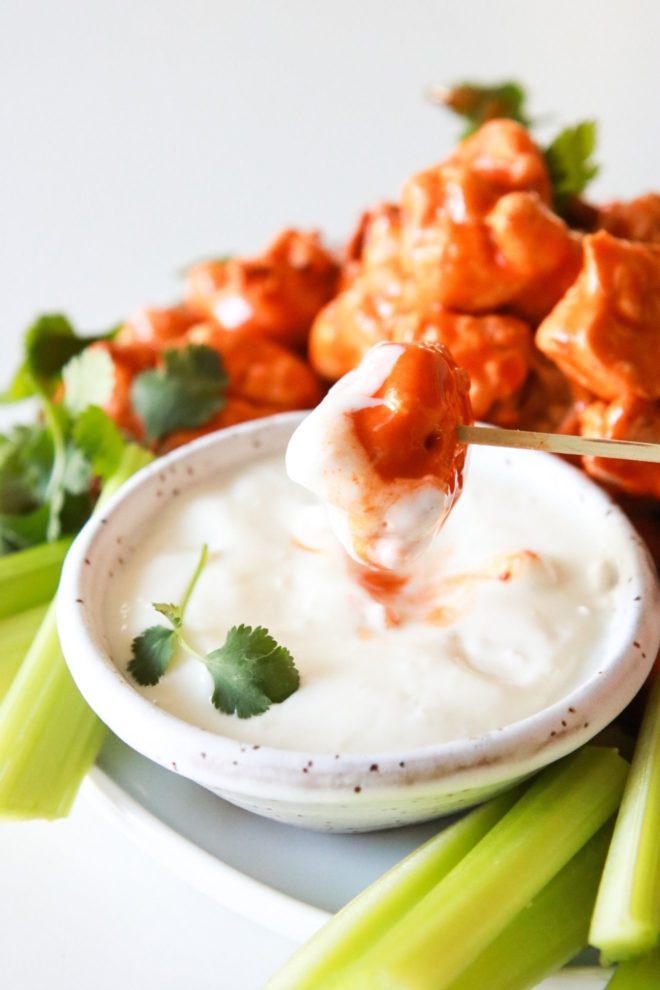 This is a side view of a plate with buffalo cauliflower florets. The plate also has a small bowl of blue cheese dip and celery sticks. One cauliflower wing is pierced with a toothpick and is being dipped into the small bowl of blue cheese dressing.