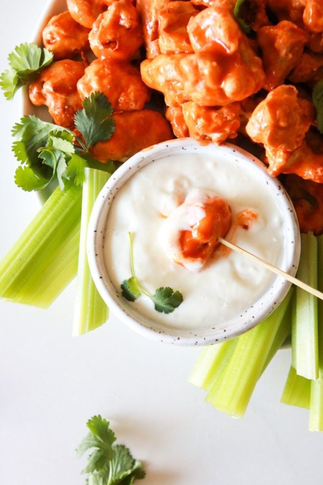 This is an overhead image of a plate with buffalo cauliflower florets. The plate also has a small bowl of blue cheese dip and celery sticks. One cauliflower wing is pierced with a toothpick and is being dipped into the small bowl of blue cheese dressing.
