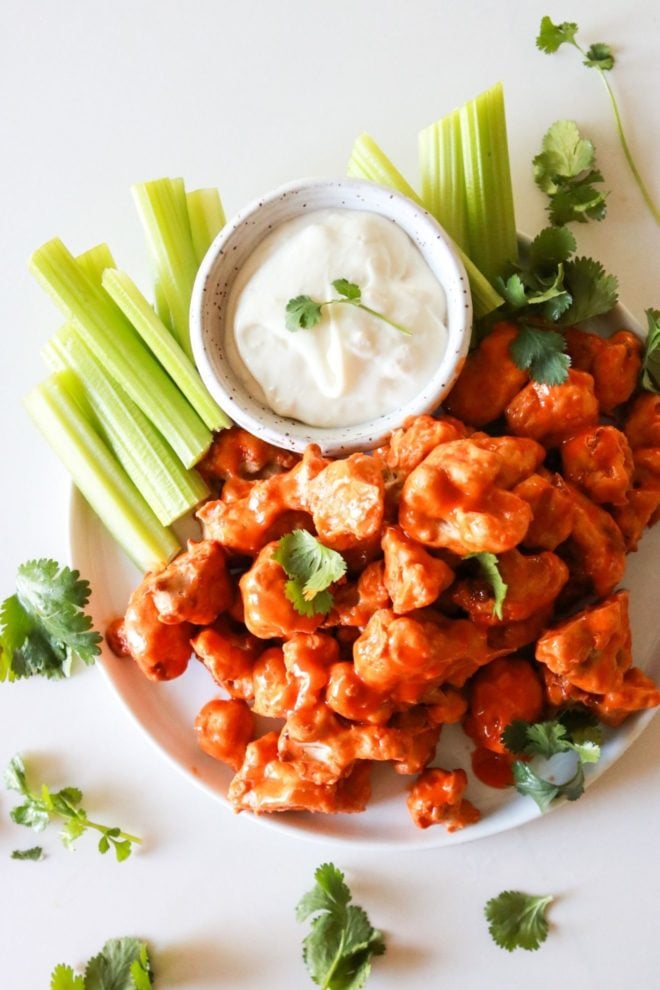 This is an overhead image of a plate with buffalo cauliflower florets. The plate also has a small bowl of blue cheese dip and celery sticks.