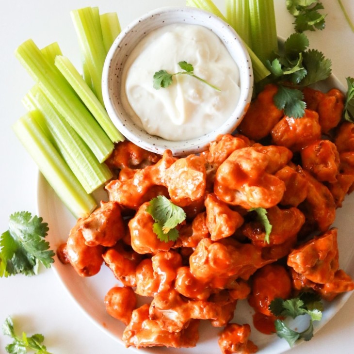 This is an overhead image of a plate with buffalo cauliflower florets. The plate also has a small bowl of blue cheese dip and celery sticks.