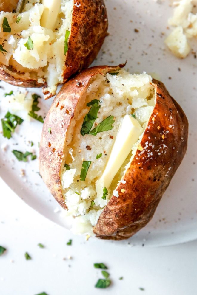 This is an overhead view of a baked potato cut in half. Inside the potato is some melted butter, freshly ground pepper and fresh herbs. The potato sits on a white plate with another potato behind it to the top left of the image.