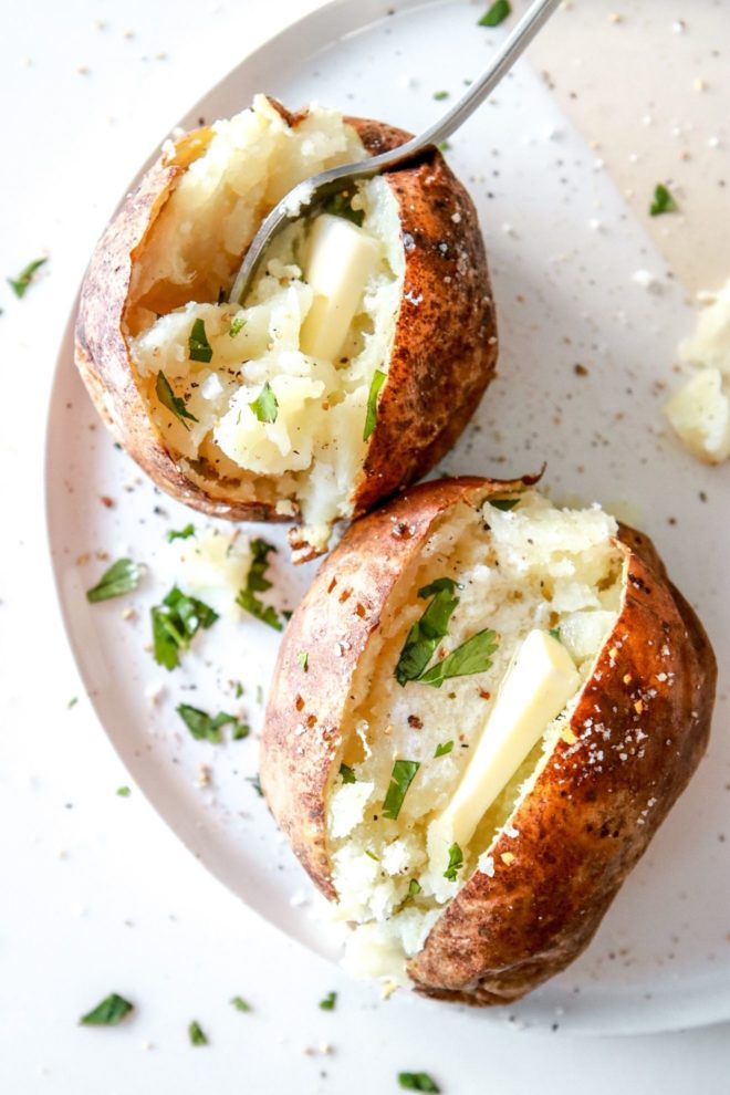 This is an overhead view of a baked potato cut in half. Inside the potato is some melted butter, freshly ground pepper and fresh herbs. The potato sits on a white plate with another potato next to the top of the image.