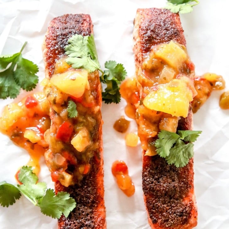 This is an overhead image of two salmon filets with jerk seasoning and mango salsa. The filets sit on a white piece of parchment paper and are garnished with fresh cilantro.