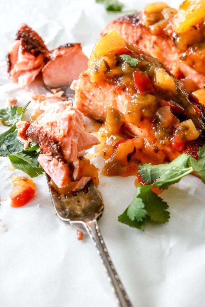 This is a side view of a fork with a bite of salmon on it. Next to the fork is a couple salmon filets with jerk seasoning and mango salsa. The salmon sits on a white piece of parchment paper and has fresh cilantro around the filets.