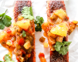 This is an overhead image of two salmon filets with jerk seasoning and mango salsa. The filets sit on a white piece of parchment paper and are garnished with fresh cilantro. Text overlay reads "jerk salmon made in the air fryer."