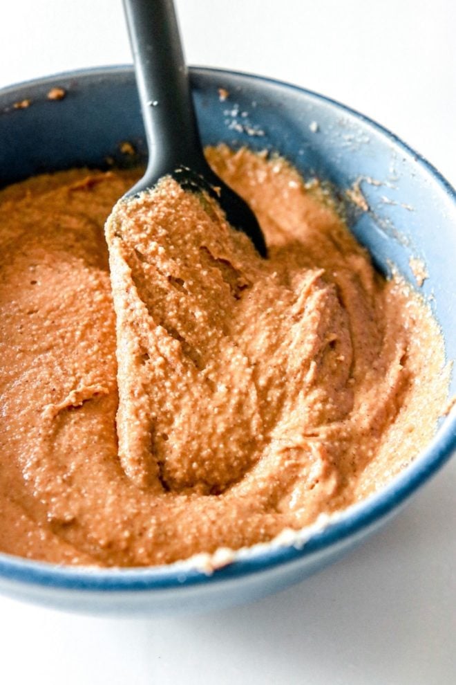 This is an image looking into a blue bowl with a nut butter mixture in it. A spatula is in the mixture and leaning against the bowl. The bowl sits on a white counter.