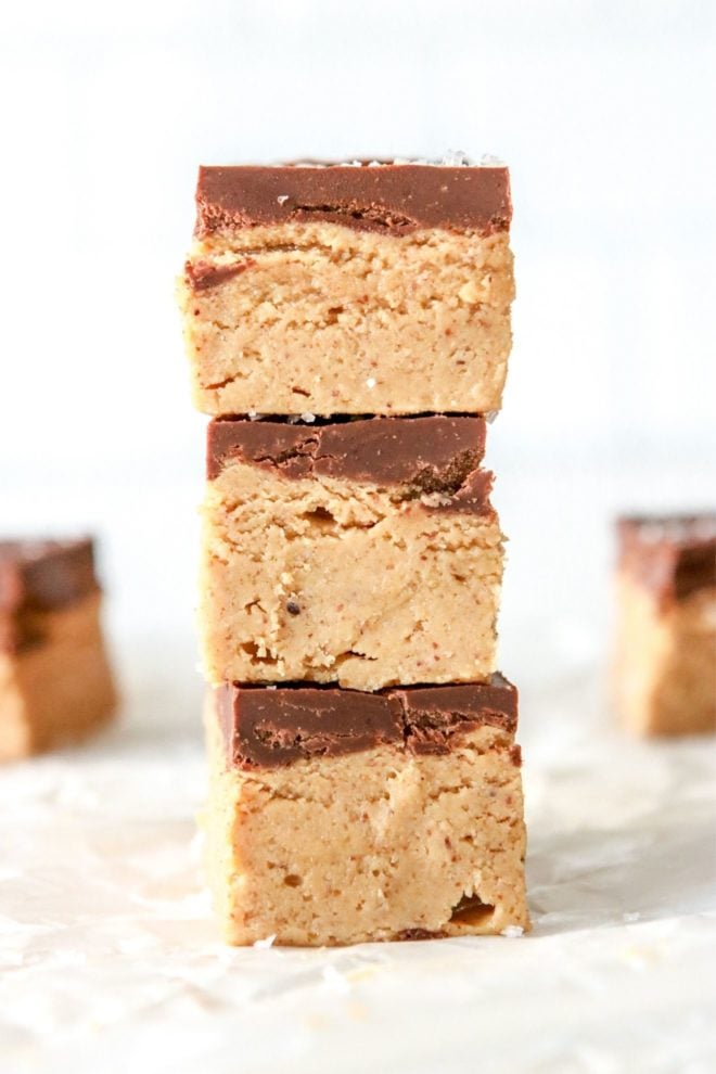 This is a side view of a stack of three chocolate almond butter bars. The stack sits on a white piece of parchment paper with a white background. More bars are blurred in the background.