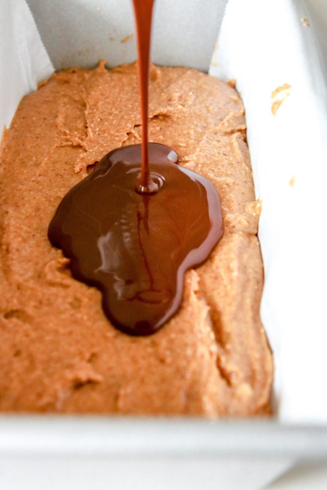 This is a close up side image of chocolate being poured onto a nut butter layer. The nut butter layer is in a pan with white parchment paper.