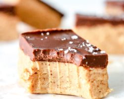 This is a side view of a chocolate almond butter bar with a bite taken out of it. The bar sits on a white piece of parchment paper and is sprinkled with salt. More bars are blurred in the background.